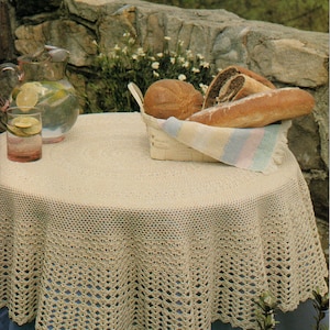 May include: A round, cream-colored crocheted tablecloth with a scalloped edge covers a table. A basket of bread and a pitcher of water with lemon slices sit on the table. The background includes a stone wall and greenery.
