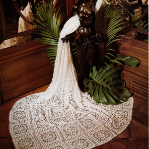 May include: A bronze statue of a woman draped with a cream-colored, intricately crocheted shawl. The shawl features a repeating floral pattern and cascades onto a terracotta tile floor. Green tropical leaves and a wooden cabinet are in the background.