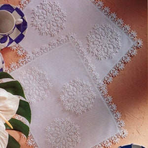 Puede incluir: Manteles individuales blancos de ganchillo con intrincados diseños florales y bordes festoneados. Los manteles individuales cuadrados presentan un diseño central rodeado por un delicado borde de encaje. Una taza y un platillo son visibles en el fondo.