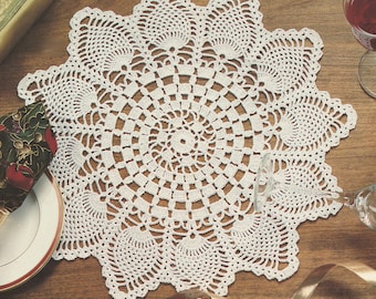 Op de afbeelding: Een witte, gehaakte doily met een cirkelvormig ontwerp en geschulpte randen. De doily is geplaatst op een houten oppervlak, met een bord, servet en wijnglas zichtbaar op de achtergrond. Het ingewikkelde kantachtige patroon is zichtbaar.