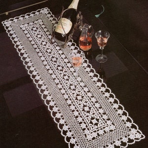 Op de afbeelding: Een witte gehaakte tafelloper met een delicaat bloemenpatroon. De loper ligt op een zwart oppervlak met een fles champagne en twee glazen wijn.