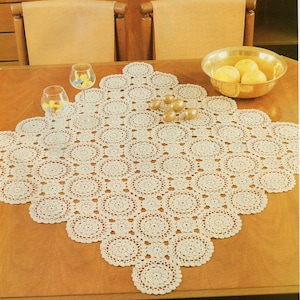 May include: A white crocheted table runner with a circular pattern. The runner is on a wooden table with two chairs and a bowl of fruit.