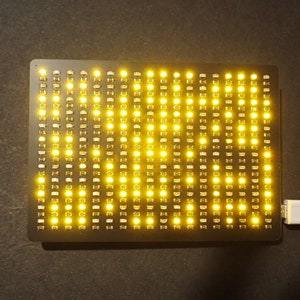 May include: A rectangular black LED matrix display with numerous yellow lights illuminated. A white USB cable is connected to the display. The lights are arranged in a grid pattern, creating a bright, eye-catching visual.