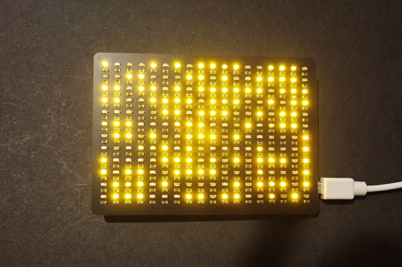 May include: A rectangular black LED matrix display with numerous yellow lights illuminated. A white USB cable is connected to the display. The lights are arranged in a grid pattern, creating a bright, eye-catching visual.