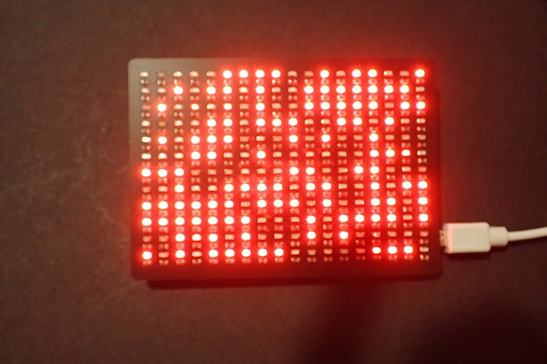 K&ouml;nnte beinhalten: Eine schwarze rechteckige Platine mit einem Raster aus roten LEDs. Die Platine ist mit einem wei&szlig;en USB-Kabel verbunden.