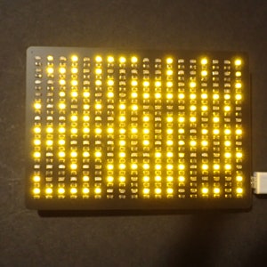May include: A rectangular black LED matrix display with numerous yellow lights illuminated. A white USB cable is connected to the display. The device is on a dark surface.