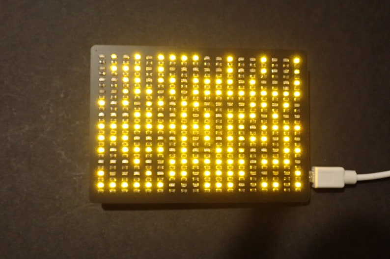 May include: A rectangular black LED matrix display with numerous yellow lights illuminated. A white USB cable is connected to the display. The device is on a dark surface.