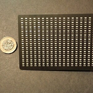 May include: A black rectangular electronic component with numerous small, white, semi-circular elements arranged in a grid pattern. A one-pound coin is next to the component for scale, showing the detail of the coin's design.