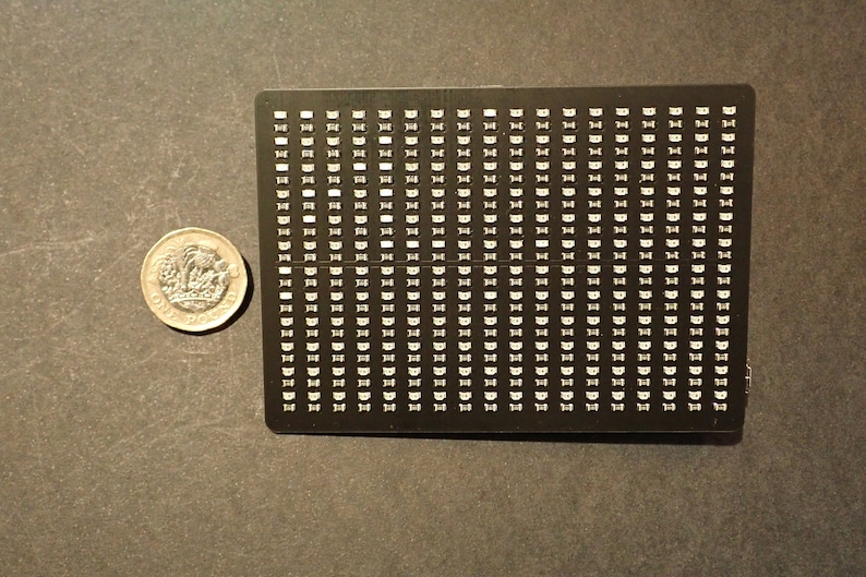 May include: A black rectangular electronic component with numerous small, white, semi-circular elements arranged in a grid pattern. A one-pound coin is next to the component for scale, showing the detail of the coin's design.