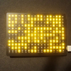 Puede incluir: Una pantalla de matriz LED negra rectangular con numerosas luces amarillas encendidas. Un cable USB blanco está conectado al dispositivo. La pantalla está sobre una superficie oscura.