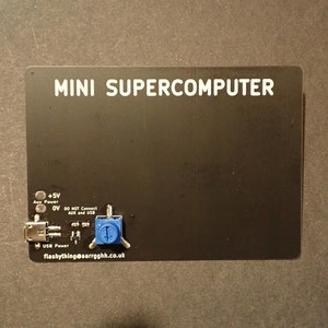 May include: A black circuit board labeled "MINI SUPERCOMPUTER" with white text. It features a USB port, a blue potentiometer, and text indicating power and connection details. The board is on a dark surface.