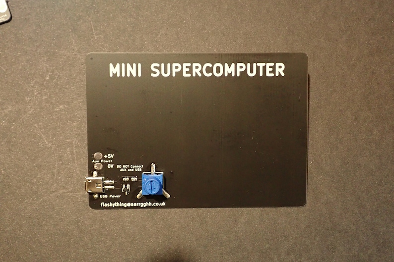 May include: A black circuit board labeled "MINI SUPERCOMPUTER" with white text. It features a USB port, a blue potentiometer, and text indicating power and connection details. The board is on a dark surface.