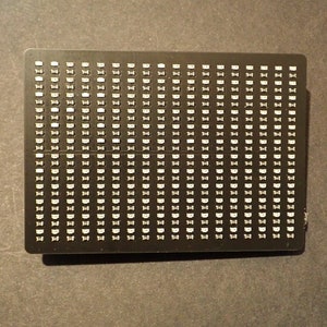 May include: A black rectangular electronic component with numerous small, white, rectangular lights arranged in a grid pattern. The component is centered and appears to be a circuit board or similar device.