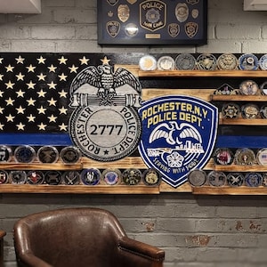 May include: A wooden wall display featuring a stylized American flag with a thin blue line, integrated shelves holding circular challenge coins, and two large circular emblems for the Rochester, N.Y. Police Dept. with the number 2777.