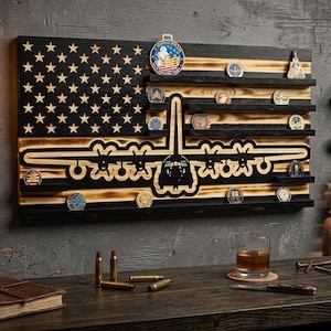 May include: A rectangular wooden wall display featuring a charred black and natural wood American flag design. The piece includes horizontal shelves for challenge coins and a central cutout silhouette of a four-engine military transport aircraft.