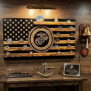 May include: A wall display featuring a wooden US Marine Corps flag with coin shelves, a framed dress uniform, a brass bell, a USMC recruitment poster, rank insignia, a commemorative plaque, a knife in a display case, and a sextant on a table.
