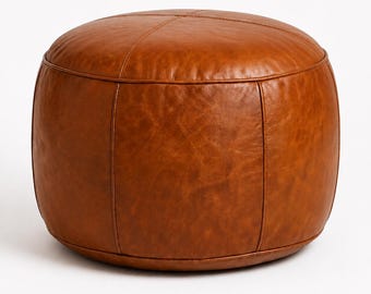 May include: A round, cognac-colored leather pouf featuring a smooth, slightly textured surface with visible vertical and horizontal stitched seams that divide the top and sides into distinct, symmetrical panels, creating a structured, cylindrical shape.