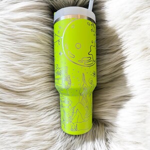 May include: A neon green insulated tumbler with a white lid and straw. The tumbler features a silver etched design of a full moon, stars, and a camping scene. The tumbler is designed to keep drinks cold or hot.