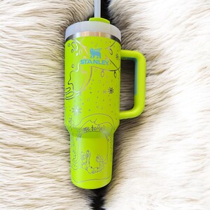 May include: A lime green Stanley tumbler with a handle and a straw. The tumbler has a silver lid and is decorated with silver illustrations of a Christmas tree, lights, and the words "Welcome to Winter". The Stanley logo is visible.