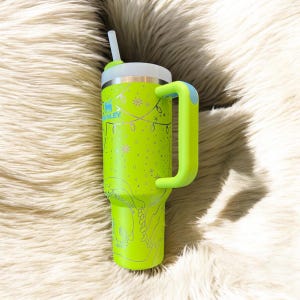 May include: A neon green insulated tumbler with a white lid and a matching handle. The tumbler features a festive design with snowflakes, string lights, and the brand name "Stanley". Includes a white straw.
