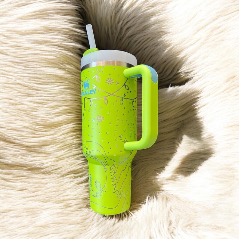 May include: A neon green insulated tumbler with a white lid and a matching handle. The tumbler features a festive design with snowflakes, string lights, and the brand name "Stanley". Includes a white straw.