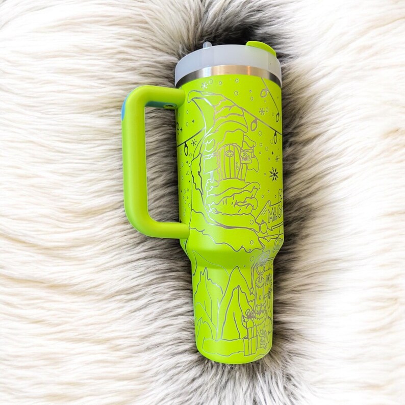 May include: A neon green travel mug with a silver lid and handle. The mug features a detailed silver engraving of a winter scene with a gnome, snowflakes, and string lights. The handle is a bright green color.