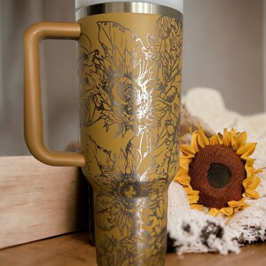 May include: Mustard-colored insulated tumbler with a handle, featuring a silver sunflower design. The tumbler has a white top and a matching mustard-colored handle. A decorative sunflower and a cream-colored blanket are also in the image.