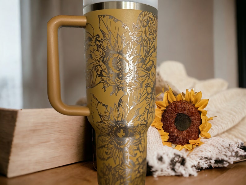 May include: Mustard-colored insulated tumbler with a handle, featuring a silver sunflower design. The tumbler has a white top and a matching mustard-colored handle. A decorative sunflower and a cream-colored blanket are also in the image.