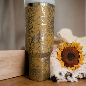 May include: A stainless steel tumbler with a mustard yellow finish and a silver floral design. A decorative sunflower and a cream-colored blanket are next to the tumbler. The tumbler is approximately 10 inches tall.