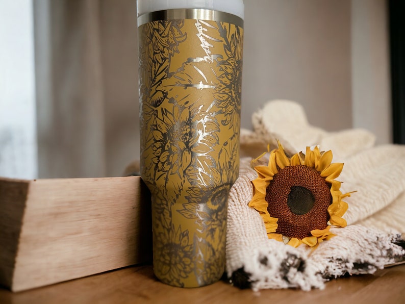 May include: A stainless steel tumbler with a mustard yellow finish and a silver floral design. A decorative sunflower and a cream-colored blanket are next to the tumbler. The tumbler is approximately 10 inches tall.