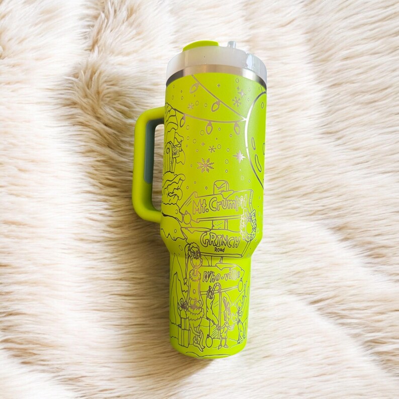 May include: A lime green travel tumbler with a handle, featuring a silver lid and a silver rim. The tumbler is decorated with a Grinch-themed design, including the text "Mt. Crumpit Road" and "Who-ville".