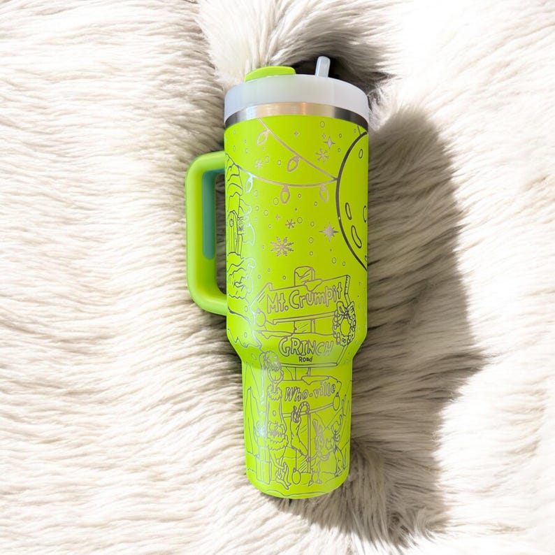 May include: A neon green insulated tumbler with a handle and a white lid. The tumbler features etched illustrations and text, including "Mt. Crumpit" and "Grinch Road". A light blue accent is on the handle.
