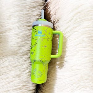 May include: A neon green Stanley tumbler with a handle and straw. The tumbler features a silver rim and a light green handle. The design includes the word "Stanley" in blue, along with a festive illustration. The tumbler is designed to hold cold beverages.