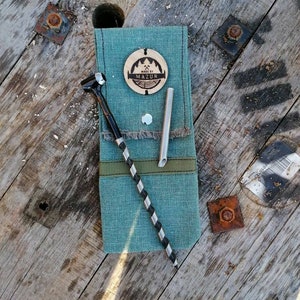 Puede incluir: Una bolsa de tela turquesa con una etiqueta de madera marrón que dice "Made by Mazur" y un taladro negro y blanco y un tubo de metal plateado en su interior.