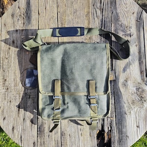 Puede incluir: Bolso mensajero de lona verde con correa para el hombro y cierre de solapa. El bolso tiene dos correas ajustables con hebillas metálicas y un reposahombros acolchado. El bolso mide aproximadamente 30 cm de ancho y 25 cm de alto.