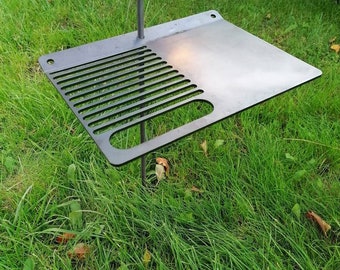May include: A black metal grilling grate with a raised edge and a handle. The grate is supported by a metal rod that is inserted into the ground.