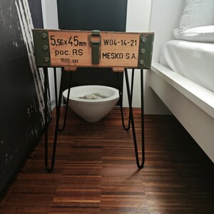 May include: A wooden crate with black metal hairpin legs. The crate is painted green and has the text "5.56x45mm poc. RS 1600 SZT W04-14-21 MESKO S.A." printed on it. The crate is sitting on a wooden floor.