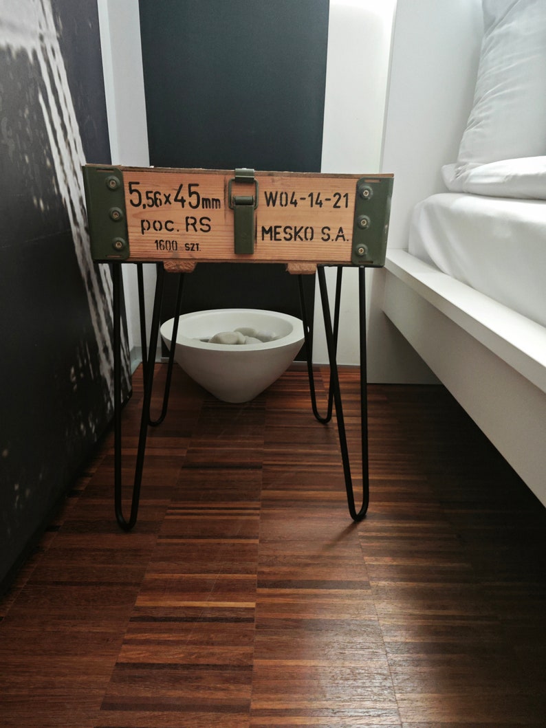 May include: A wooden crate with black metal hairpin legs. The crate is painted green and has the text "5.56x45mm poc. RS 1600 SZT W04-14-21 MESKO S.A." printed on it. The crate is sitting on a wooden floor.
