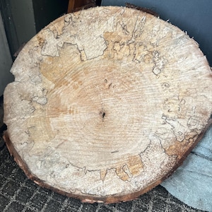 May include: A cross-section of a tree trunk, showcasing the natural wood grain and patterns. The light brown wood has darker, intricate markings and a rough, textured bark edge. The circular shape reveals the tree's growth rings.