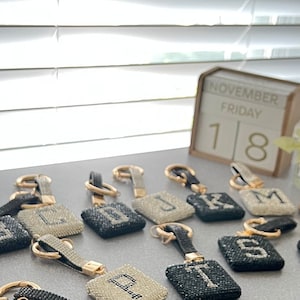 May include: A collection of 18 black and white rhinestone letter keychains with gold rings. The keychains are arranged in a semi-circle on a white surface. The letters are A, B, C, D, J, K, L, M, N, P, S, T, and Z.