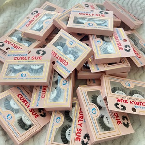 May include: A pile of pink and gold boxes containing false eyelashes. The boxes are labelled "Curly Sue" and "Hot Roller Lash". Some of the boxes are open, revealing the eyelashes inside.
