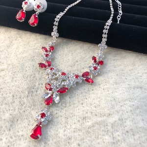 May include: A silver necklace with a teardrop-shaped pendant and red crystal accents. The necklace is paired with matching earrings.