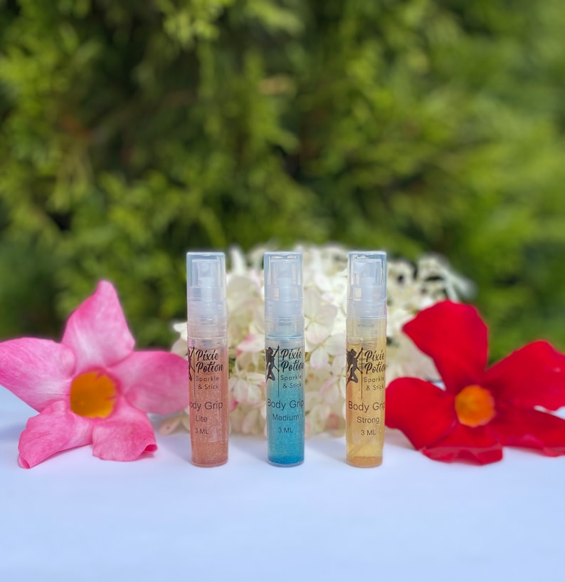 Może przedstawiać: Trzy małe butelki z rozpylaczem "Pixie Potion" Body Grip, oznaczone jako "Lite", "Medium" i "Strong". Butelki są prezentowane z r&oacute;żowymi i czerwonymi kwiatami na zielonym tle. Każda butelka zawiera 3 ml produktu.