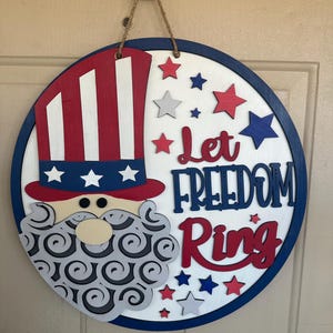 May include: A round wooden door hanger with a blue and white border. The center features a red, white, and blue Uncle Sam hat with a gray beard and the text "Let Freedom Ring" in red and blue.