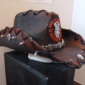 May include: A brown and black cowboy hat with a black leather band and silver studs. The hat has a patch on the front that says "Sons of Anarchy Est. Redwood Original".