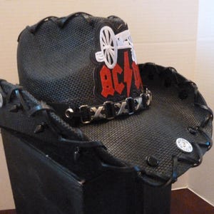 Op de afbeelding: Zwarte cowboyhoed met een zwart-wit AC/DC-logo en een zilveren kettingdetail.