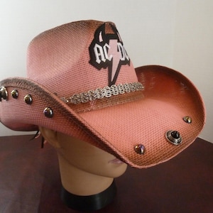 May include: Pink cowboy hat with the AC/DC logo. The hat features a woven texture, a decorative band, and metal studs around the brim. The hat is displayed on a mannequin head.