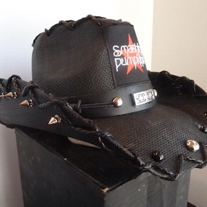 Puede incluir: Sombrero de vaquero negro con textura tejida, adornado con tachuelas y púas plateadas. El sombrero presenta una banda de cuero negro con detalles plateados y un parche con el texto "Smashing Pumpkins" y una estrella roja.