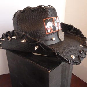 May include: Black cowboy hat with a woven texture, adorned with silver-colored studs along the brim. Features a leather band with silver studs and a patch with a band logo. A decorative coin is attached to the band.