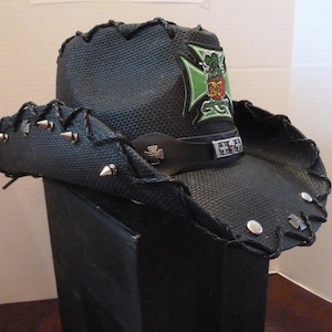 May include: A black leather cowboy hat with a green and black cross design and silver studs. The hat has a black band with a silver skull and crossbones design.